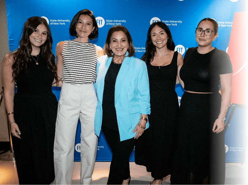 Five women posing together in front of a blue 'State University of New York' backdrop. The group includes a woman in a black dress, a woman in a black-and-white striped top and white pants, a woman in a bright blue blazer over a black outfit, a woman in a black dress, and a woman in a black crop top and long skirt.