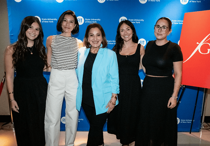 Five women posing together in front of a blue 'State University of New York' backdrop. The group includes a woman in a black dress, a woman in a black-and-white striped top and white pants, a woman in a bright blue blazer over a black outfit, a woman in a black dress, and a woman in a black crop top and long skirt.