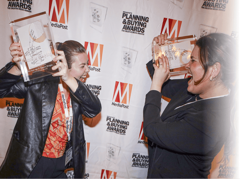 Two women laughing joyfully while holding up clear acrylic awards at MediaPost's Planning & Buying Awards, standing in front of a step-and-repeat banner covered in MediaPost logos.
